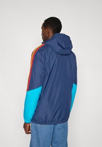 Marineblauwe windbreakerjas met capuchon; heeft rode en gele strepen, turquoise accenten op de mouwen, elastische manchetten en een casual pasvorm.