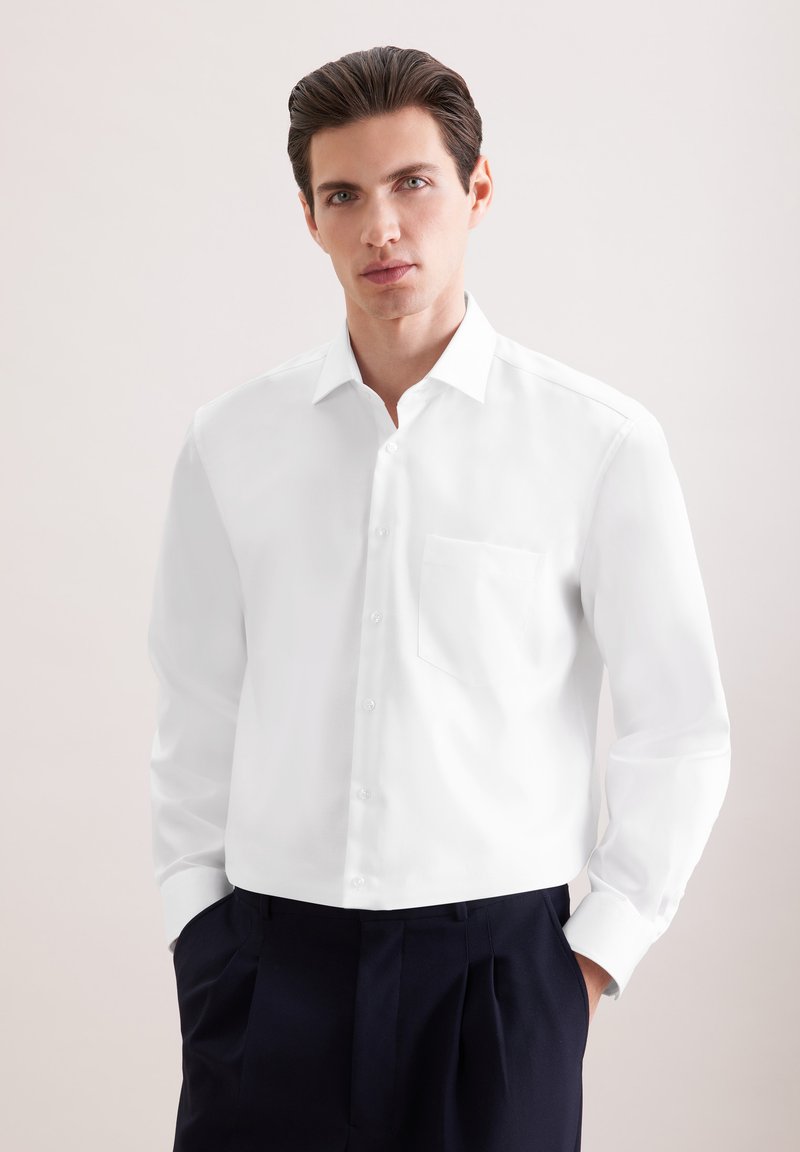 Homme aux cheveux foncés portant une chemise blanche boutonnée et un pantalon foncé, debout les mains dans les poches devant un fond clair uni.