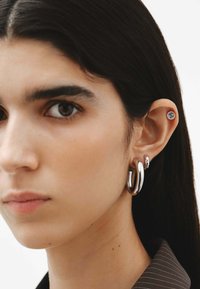 Pendientes de aro plateados y un pequeño pendiente de piedra preciosa, llevados en la oreja de una modelo, con piel suave y cabello oscuro, con un fondo mínimo.