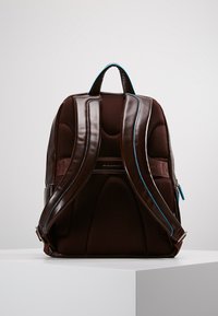 Mochila de cuero marrón con respaldo de malla acolchada, acentos azules, correas de hombro ergonómicas y un asa superior para llevar.