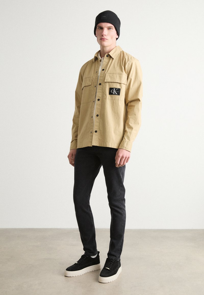 Beżowa koszula z guzikami, z dwoma kieszeniami na piersi i czarną łatą z logo, zestawiona z dopasowanymi czarnymi dżinsami i czarnymi sneakersami na platformie. Na głowie czarna czapka beanie.