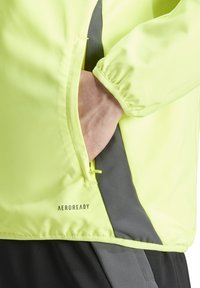 Veste jaune vif avec une texture lisse, comportant une poche zippée et des accents gris sur le côté. Logo "AEROREADY" affiché.