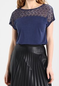 Námořnická modrá blůza s krajkovou aplikací na ramenou; krátké rukávy, kulatý výstřih. V kombinaci s černou skládací sukní. Měkká tkanina, hladká textura.
