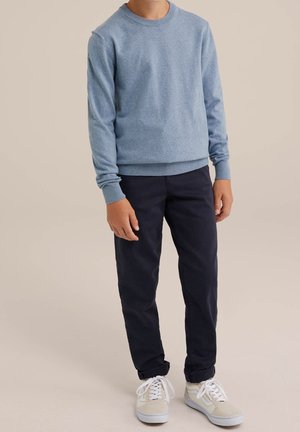 Enfant portant un pull bleu clair à manches longues, un pantalon sombre et des baskets crème avec des lacets blancs, debout devant un fond uni.