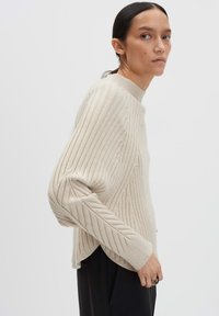 Donna in un maglione a costine color crema con maniche a palloncino e colletto alto, che indossa pantaloni neri, in piedi davanti a uno sfondo chiaro e semplice.