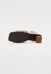 Sandale dorée à lanières avec une semelle noire texturée, un talon bloc et des détails tressés. Dispose d'une sangle de cheville ajustable pour un meilleur maintien.