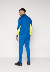 Μπλε αθλητική φόρμα με κίτρινες λεπτομέρειες. Το μπουφάν έχει ψηλό γιακά και εφαρμοστά μανίκια με τελείωμα. Το παντελόνι είναι στενό με ριμπ μπατζάκια.