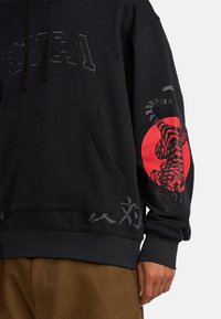 Schwarzer Hoodie mit einer Fronttasche, versehen mit dem "RVCA"-Logo, einem roten Tiger-Grafik auf dem Ärmel und dekorativem grauem Text entlang des Saums.