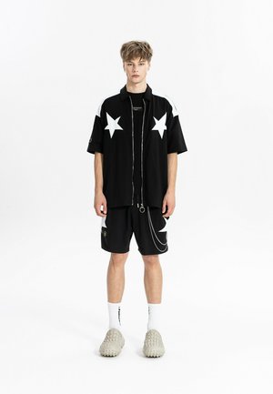 Persona joven que lleva una camiseta negra de manga corta y pantalones cortos con estrellas blancas, calcetines blancos y zapatos beige texturizados sin cordones, de pie sobre un fondo blanco.