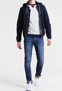 Giacca navy con zip sopra un felpa grigia, abbinata a jeans blu e sneakers bianche. Design casual con cappuccio e texture in contrasto.