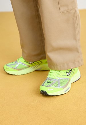 Pieds portant des baskets Nike vert néon et argentées associées à un pantalon beige à jambes larges sur un sol jaune moutarde.