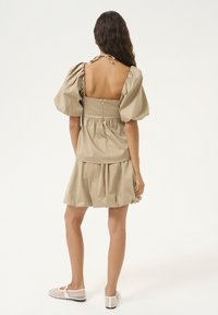 Abito color khaki con maniche a palloncino, schiena quadrata, vita arricciata e gonna a strati. Indossato con scarpe flat trasparenti. Tessuto di cotone morbido.