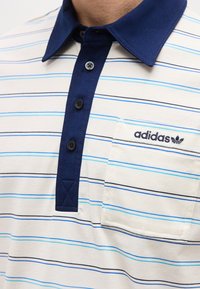 Λευκό πόλο μπλουζάκι με λεπτές μπλε ρίγες, σκούρο μπλε γιακά και ταινία με κουμπιά, τσέπη στο στήθος με το λογότυπο Adidas και το σύμβολο τριφυλλιού.