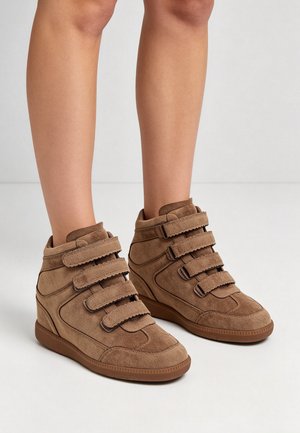 Baskets compensées en daim marron avec quatre sangles auto-agrippantes réglables, portées sur des jambes nues sur un fond clair uni.