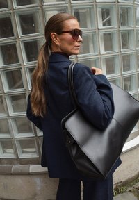 Ženska z dolgimi rjavimi lasmi, spet v čop, nosi sončna očala in temno modro blazer jakno, nosi veliko črno usnjeno torbo ob steni iz steklenih blokov.