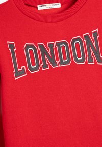 Sudadera roja de tejido suave con "LONDRES" en letras negras y blancas en negrita, cuello redondo y puños acanalados.