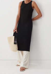 Svart ärmlös klänning med ribbad textur, i kombination med krämfärgade vida byxor. Håller en vävd beige väska med svarta remmar. Platta sandaler synliga.