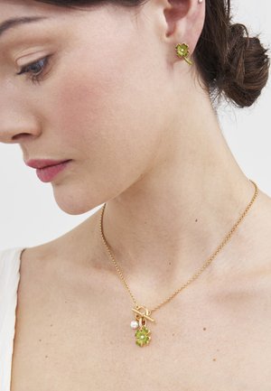 Žena nosi zlatnu ogrlicu sa privjeskom zelenim djetelinom s četiri lista i biserom, uz uhu naušnice s istim motivom djeteline, vidljiv bijeli vrh.