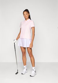 Ροζ πόλο μπλούζα και λευκή φούστα με διακριτικό σχέδιο. Λευκά παπούτσια γκολφ και γάντι. Πόζα με μπαστούνι του γκολφ μπροστά από ουδέτερο φόντο.