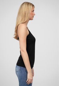 Svart tank topp laget av bomull, med tynne stropper og en tettsittende design. Kombinert med lyseblå jeans, vist fra siden.