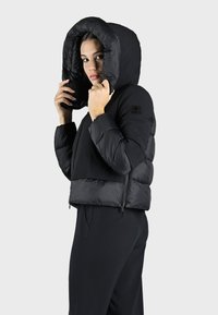 Zwarte gewatteerde jas met een oversized capuchon, cropped ontwerp, ritssluiting en textuurafwerking. Accenten met een merklogo op de mouw.