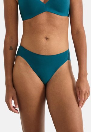 Kobieta ubrana w spodnie od bikini w kolorze morskim oraz pasujący top, pokazująca środkową część tułowia i górne uda, z małym geometrycznym tatuażem na lewym przedramieniu.