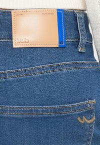 Jeans in denim blu con etichetta in pelle marrone chiaro che presenta dettagli cuciti e un accento blu. Cuciture evidenti attorno alle tasche e alla vita.