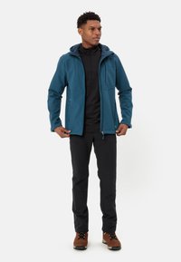 Turkusowa kurtka softshell z kapturem, przednimi kieszeniami i czarnym zapinanym na zamek pod spodem, w połączeniu z czarnymi spodniami i brązowymi butami trekkingowymi.