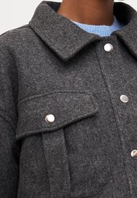 Szara wełniana kurtka z kołnierzem, srebrnymi zatrzaskami, kieszenią na piersi i miękką fakturą. Częściowo widoczna jest niebieska podszewka.