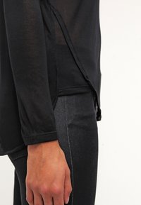 Ruka odpočívající vedle černé dlouhé košile s bočním rozparkem, v kombinaci s tmavými upnutými kalhotami na jednoduchém světlém pozadí.