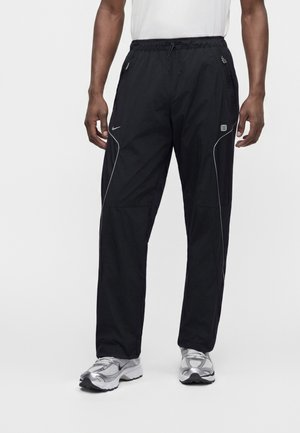 Mandlig model iført sorte Nike træningsbukser med hvide reflekterende striber og grå løbesko, stående mod en ensfarvet hvid baggrund.