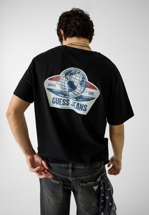 Uomo che indossa una t-shirt nera Guess Jeans con logo del globo e scritta sul retro, jeans scuri con una bandana nera infilata in tasca.