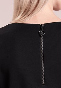 Schwarzes Kleidungsstück mit weitem Ausschnitt und sichtbarem Reißverschluss auf dem Rücken. Der Reißverschluss hat einen runden Ziehgriff und eine elegante Metalloptik.