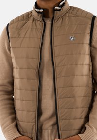Gilet rembourré beige avec un motif matelassé vertical, col haut et bordures contrastées bleu marine le long de la fermeture éclair. Porté par-dessus un pull marron clair.