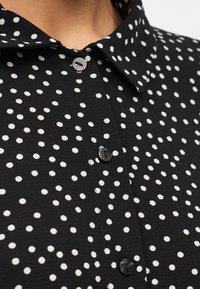 Μαύρο πουκάμισο με γιακά και ακανόνιστες λευκές πουά, κουμπωμένο, εμφανιζόμενο από τον γιακά μέχρι το μέσο του στήθους.