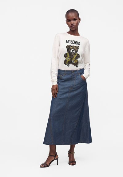 Femme portant un pull blanc Moschino avec un motif ourson, une longue jupe en jean bleue et des sandales noires à talons, debout avec une main dans la poche.
