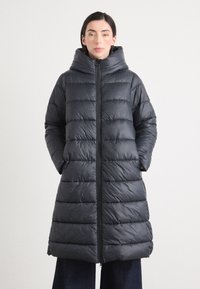 Een vrouw staat zelfverzekerd met een puffy, donkergrijze lange jas met een capuchon, handen in de zakken, tegen een neutrale achtergrond.