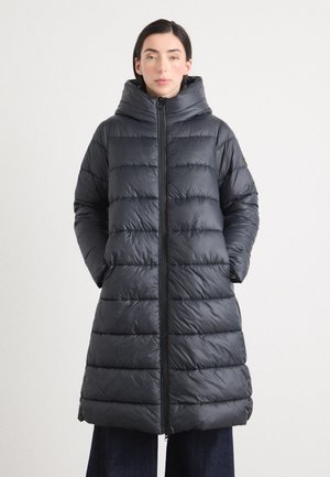 Een vrouw staat zelfverzekerd met een puffy, donkergrijze lange jas met een capuchon, handen in de zakken, tegen een neutrale achtergrond.