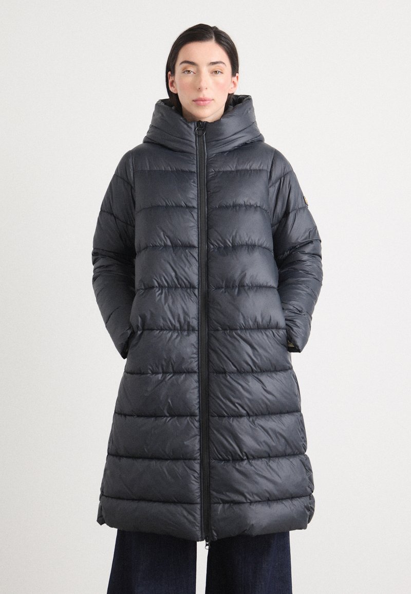 Een vrouw staat zelfverzekerd met een puffy, donkergrijze lange jas met een capuchon, handen in de zakken, tegen een neutrale achtergrond.
