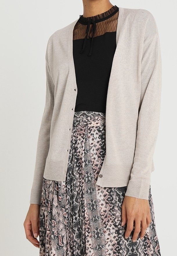 Femme portant un cardigan beige par-dessus un haut noir avec un décolleté transparent et une jupe plissée à motif peau de serpent.
