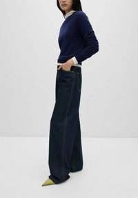 Marineblauwe trui met een ronde hals, crèmekleurige binnenlaag, wijde donkere spijkerbroek met oranje stiksel en puntige groene schoenen.