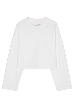 Personne portant un gilet beige sans manches boutonné sur une chemise blanche à manches longues, un pantalon blanc avec cordon et des baskets blanches épaisses, debout contre un mur blanc.