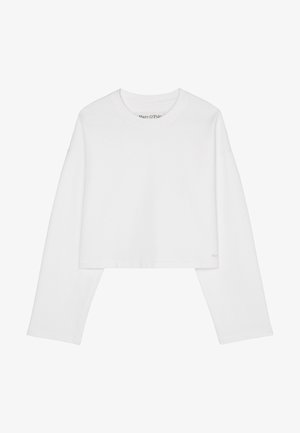 Personne portant un gilet beige sans manches boutonné sur une chemise blanche à manches longues, un pantalon blanc avec cordon et des baskets blanches épaisses, debout contre un mur blanc.