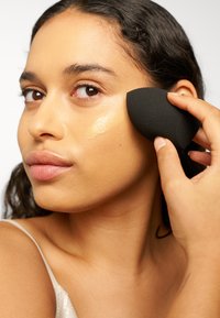 Una esponja de maquillaje negra aplicando base líquida sobre piel clara, con un enfoque en la aplicación suave y la textura natural de la piel.