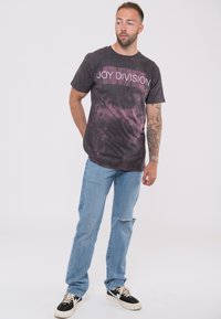 Dark gray T-shirt with a pink "JOY DIVISION" logo, paired with light blue jeans and black sneakers. The shirt has a tie-dye pattern.