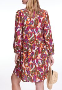 Farbenfrohes Kleid mit Paisleymuster aus leichtem Stoff, mit langen Puffärmeln und einem gestuften Saum. Ein Strohhut wird in der linken Hand gehalten.