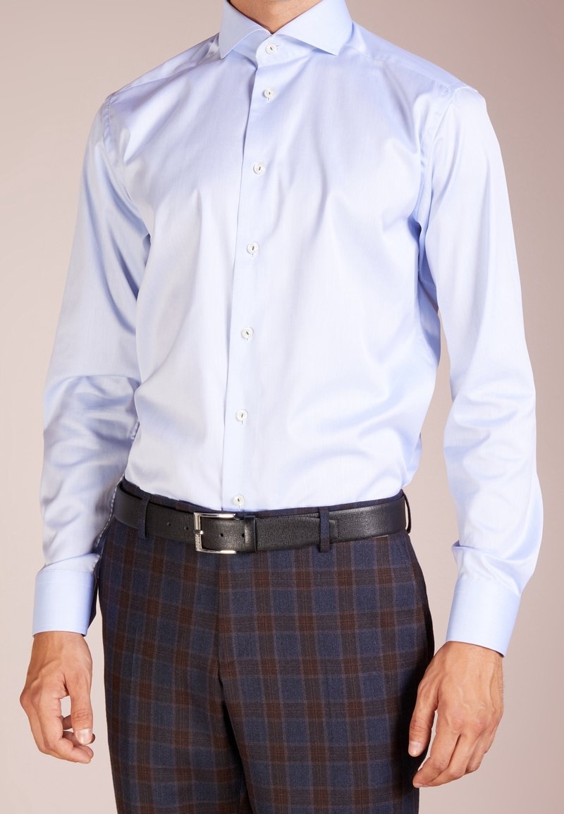 Světle modrá košile s knoflíčky a špičatým límečkem, doplněná tmavými kostkovanými kalhotami a černým opaskem. Kalhoty mají hnědé a modré tóny.