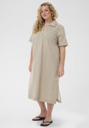 En kvinde med krøllet hår står iført en beige og hvidstribet knælængde kjole med korte ærmer og sorte flip-flops.