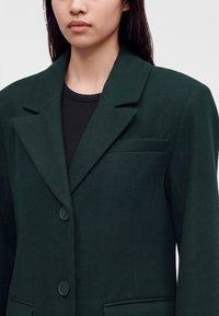 Femme portant un blazer vert foncé à revers crantés et deux boutons devant, sur un haut noir côtelé.