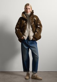 Giacca a vento marrone lucido, colletto alto, abbinata a un maglione beige chiaro a maglia a trecce, jeans blu larghi e sneakers color crema con dettagli bianchi.